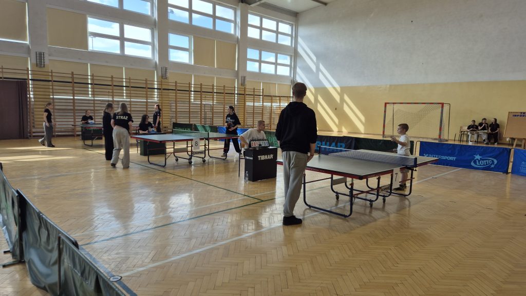 Hala sportowa z dwoma stołami do tenisa stołowego, kilkoma osobami grającymi i sędziami, jasne światło przez duże okna, drewniana podłoga, na ścianie bramka do piłki ręcznej.