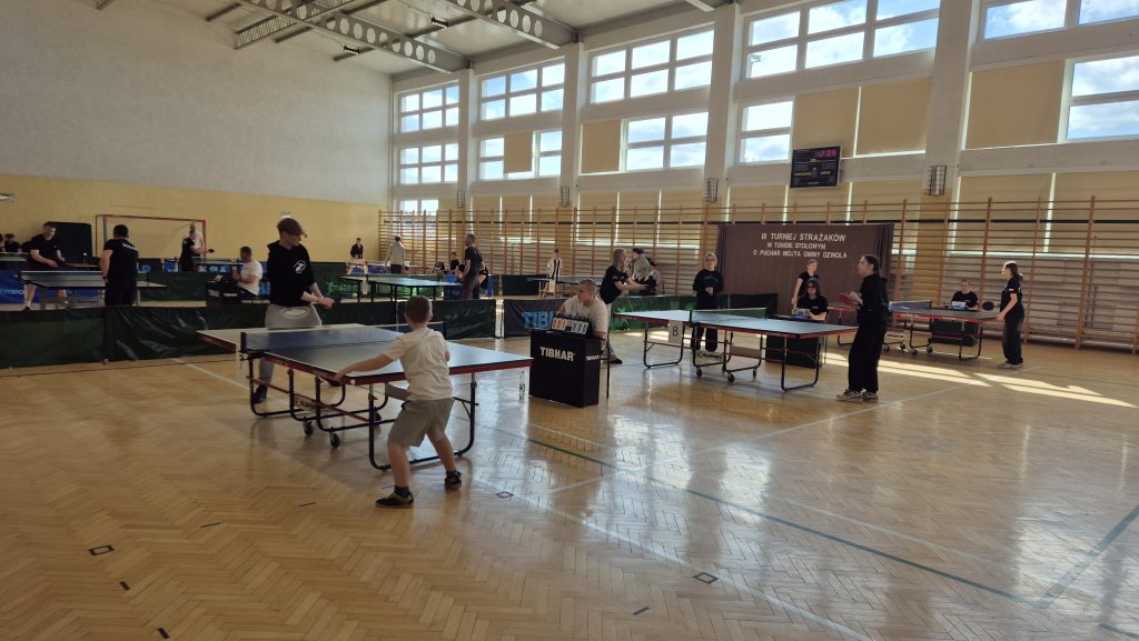 Hala sportowa z drewnianą podłogą, dużymi oknami, kilkoma stołami do tenisa stołowego i osobami grającymi lub siedzącymi przy stołach.