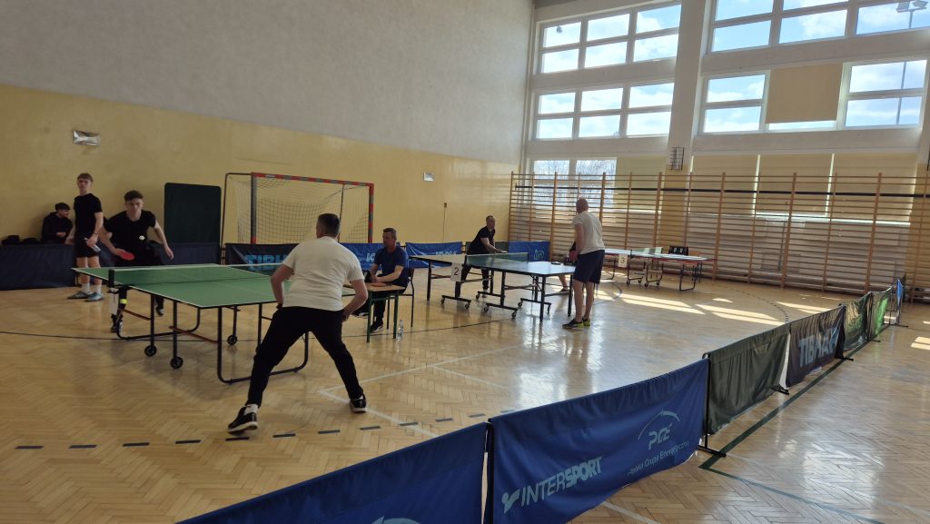 Hala sportowa z dużymi oknami, trzy stoły do tenisa stołowego, cztery osoby grające, dwie siedzące, drewniana podłoga, żółta ściana, niebieskie i zielone bandy ochronne.