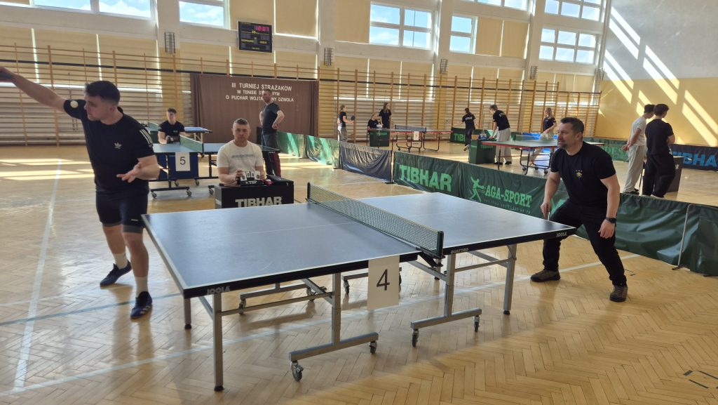 Hala sportowa z kilkoma stołami do tenisa stołowego, dwóch mężczyzn grających przy stole numer 4, sędzia z licznikiem punktów, jasne światło przez okna.