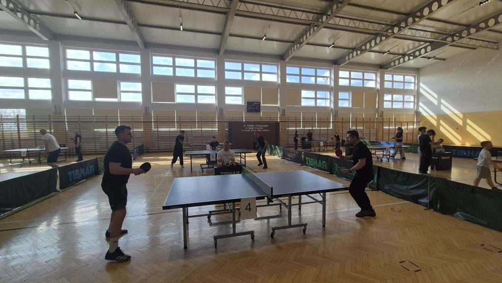 Hala sportowa z dużymi oknami, drewnianą podłogą i kilkoma stołami do tenisa stołowego, przy jednym stoi dwóch mężczyzn grających w czarnych strojach.