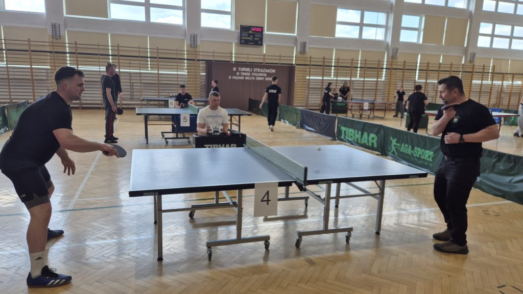 Dwóch mężczyzn w czarnych strojach gra w tenisa stołowego przy niebieskim stole z numerem 4, w tle sędzia i inni zawodnicy w jasnej, dużej hali sportowej.