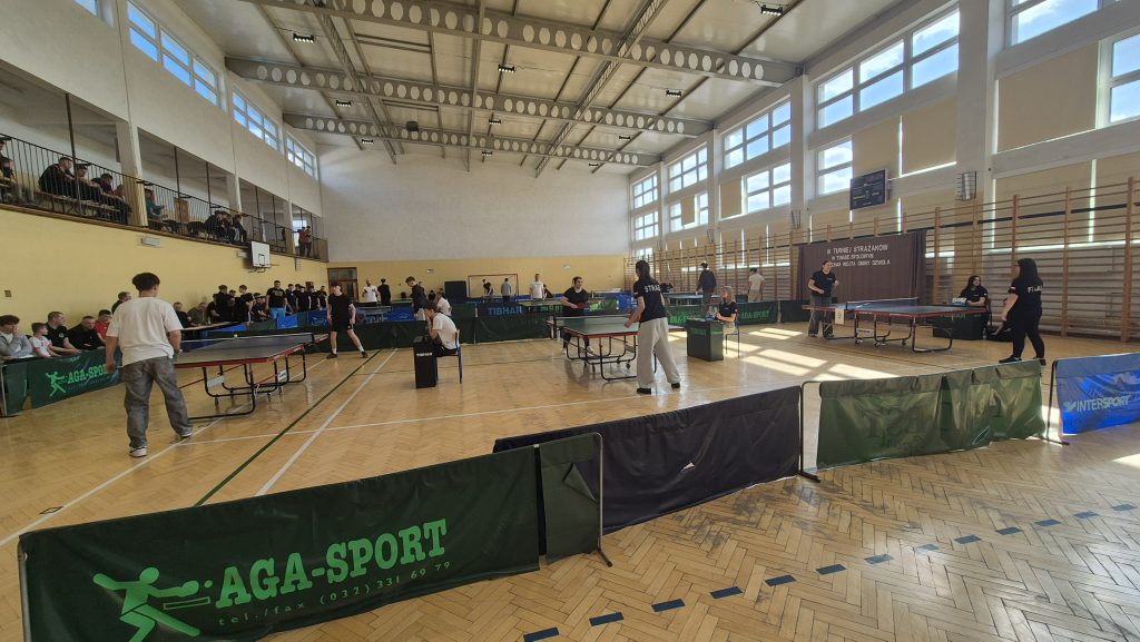 Hala sportowa z dużą liczbą osób grających i oglądających turniej tenisa stołowego, stoły do ping-ponga, drewniana podłoga, jasne okna i wysokie sufity.