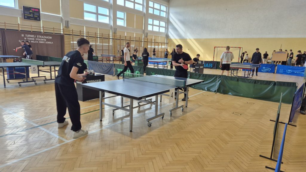 Hala sportowa z wieloma stołami do tenisa stołowego, zawodnicy w czarnych i jasnych strojach grający i obserwujący mecze, drewniana podłoga, duże okna.