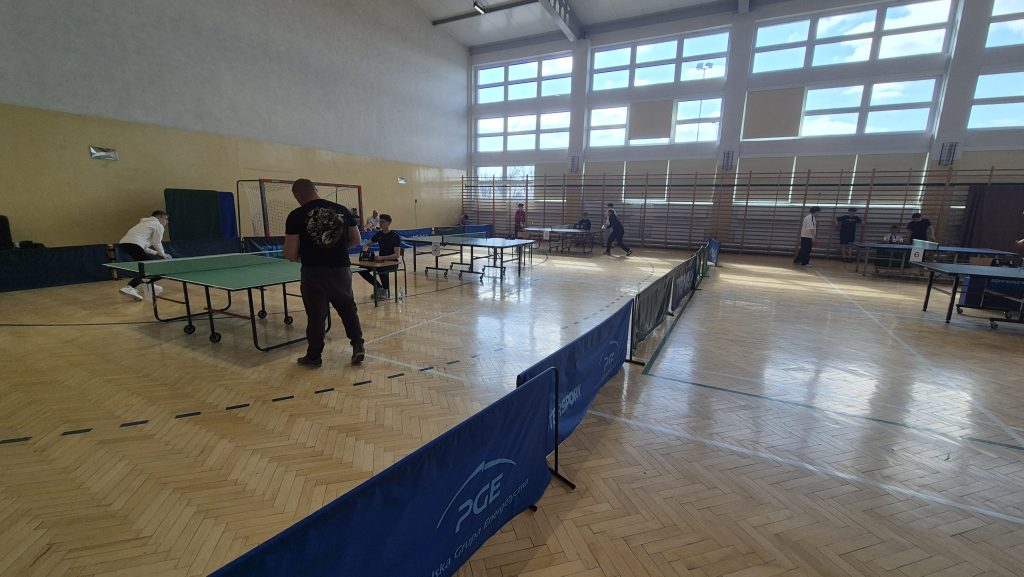 Hala sportowa z drewnianą podłogą, stoły do tenisa stołowego, kilka osób grających i obserwujących, duże okna z widokiem na niebieskie niebo.