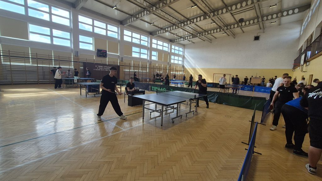 Hala sportowa z dużymi oknami, drewniana podłoga, kilka stołów do tenisa stołowego, ludzie grający i obserwujący rozgrywki, jasne oświetlenie naturalne.