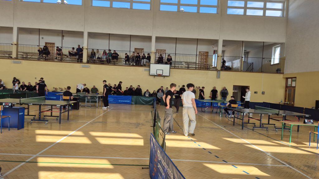 Hala sportowa z kilkoma stołami do tenisa stołowego, stojący i siedzący młodzi ludzie, żółta ściana, drewniana podłoga, górna trybuna z widzami, okna z niebieskim niebem.