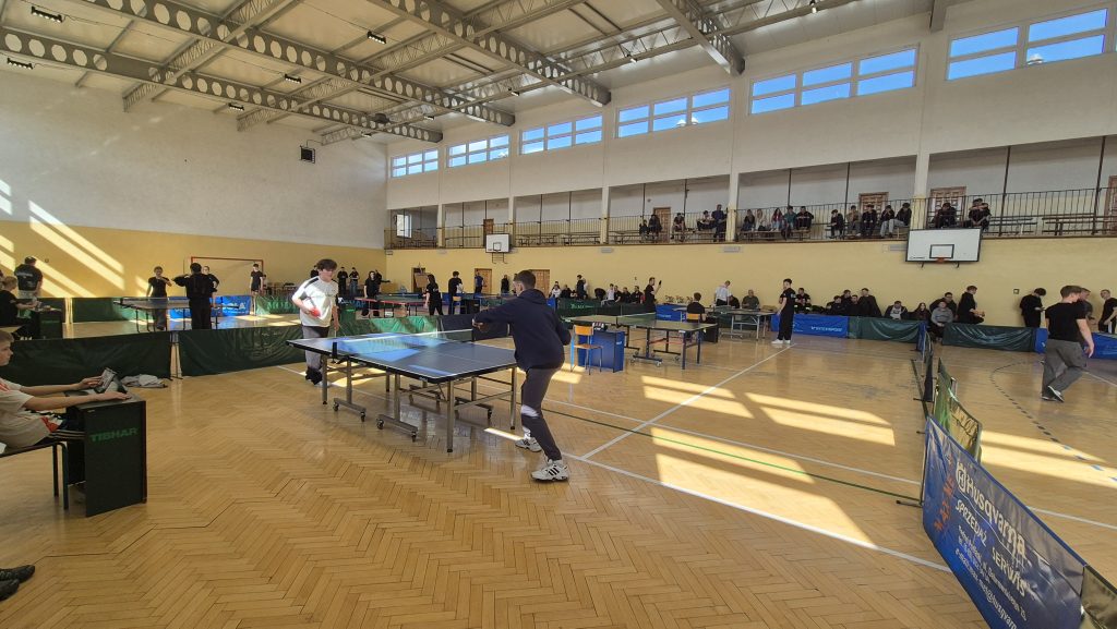 Hala sportowa z jasnym drewnianym parkietem, stoły do tenisa stołowego, zawodnicy w sportowych ubraniach, widzowie na trybunach, żółte ściany, duże okna nad nimi.