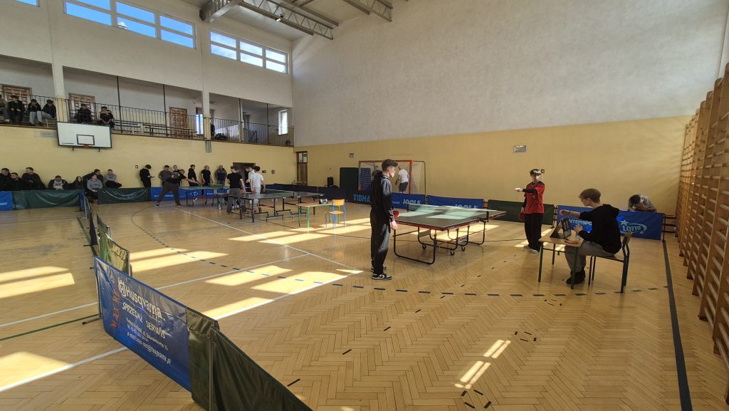 Sala gimnastyczna z jasną podłogą, stoły do tenisa stołowego, kilka osób grających i siedzących, okna na górze, ściany w kolorze białym i żółtym.