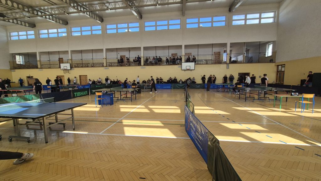 Hala sportowa z drewnianą podłogą, stoły do tenisa stołowego, ludzie stojący i siedzący wzdłuż ściany oraz na trybunach, jasne światło przez okna górne.