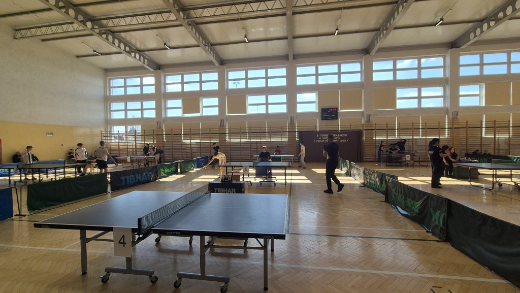 Hala sportowa z kilkoma stołami do tenisa stołowego, zawodnicy w sportowych strojach, duże okna z widokiem na niebieskie niebo, drewniana podłoga.