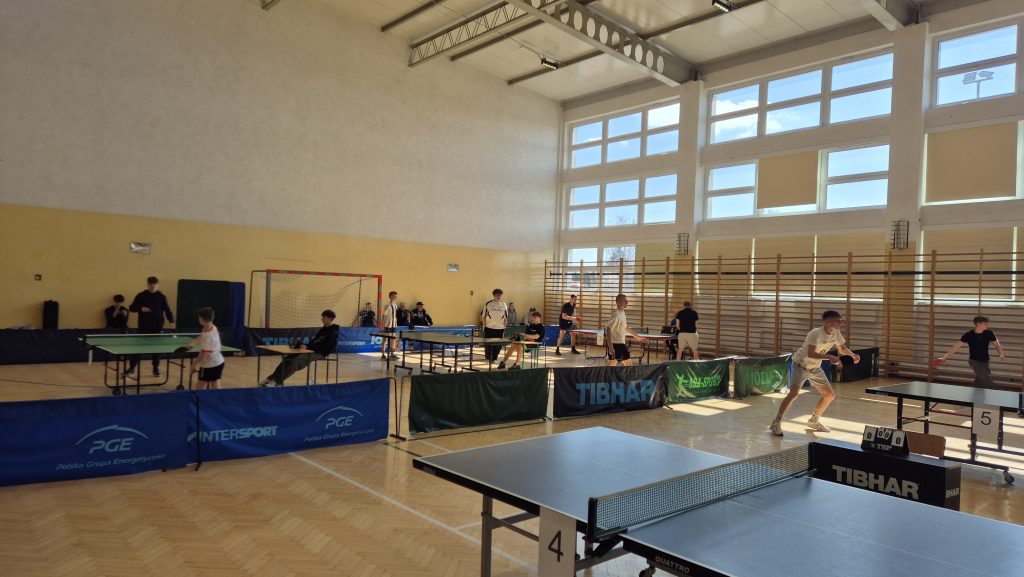 Hala sportowa z dużymi oknami, stoły do tenisa stołowego, zawodnicy w sportowych strojach grający i siedzący na krzesłach wokół stołów.