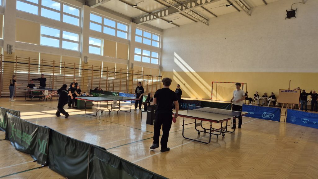 Hala sportowa z dużymi oknami, stoły do tenisa stołowego, ludzie grający i obserwujący, drewniana podłoga, siatka do piłki ręcznej na ścianie, banery reklamowe.