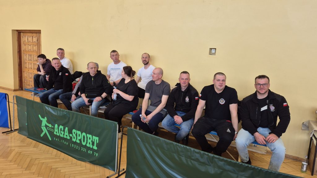 Grupa jedenastu osób, głównie mężczyzn, siedzi na ławce przy jasnej ścianie w sali sportowej, przed zielonymi barierkami z napisem AGA-SPORT.