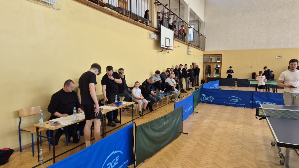 Hala sportowa z drewnianą podłogą, niebieskie i zielone bandy, ludzie stojący i siedzący na ławkach wzdłuż jasnej ściany, stoły do tenisa stołowego, kosz do koszykówki.