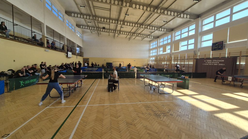 Hala sportowa z jasną podłogą, cztery stoły do tenisa stołowego, zawodnicy i sędzia, widzowie na trybunach i stojący przy ścianie, duże okna, tablica wyników, brązowa kurtyna z napisem.