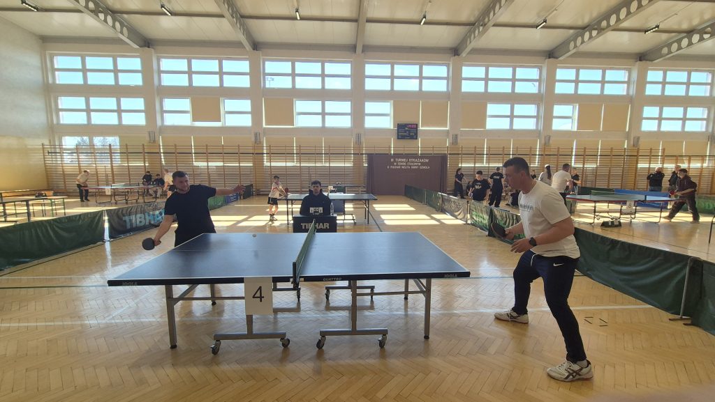 Hala sportowa z dużymi oknami, parkietem i stołami do tenisa stołowego, dwóch mężczyzn gra w tenisa, sędzia przy stole z licznikiem punktów.