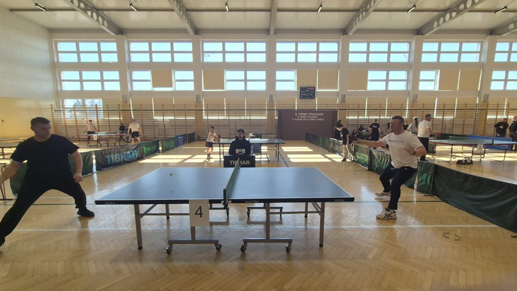 Hala sportowa z dużymi oknami, drewnianą podłogą, stołem do tenisa stołowego z siatką i zawodnikami w trakcie gry, sędzia przy stole.