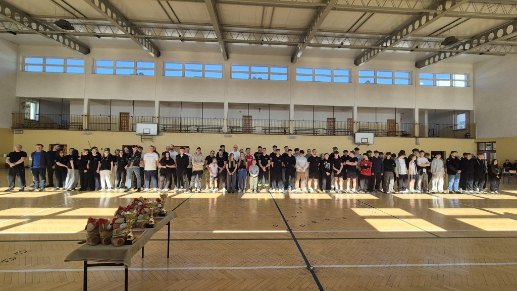 Wielka grupa ludzi w sportowej hali na drewnianej podłodze, w tle żółte ściany, okna i kosze do koszykówki, na pierwszym planie stół z pucharami i pluszowymi misiami.