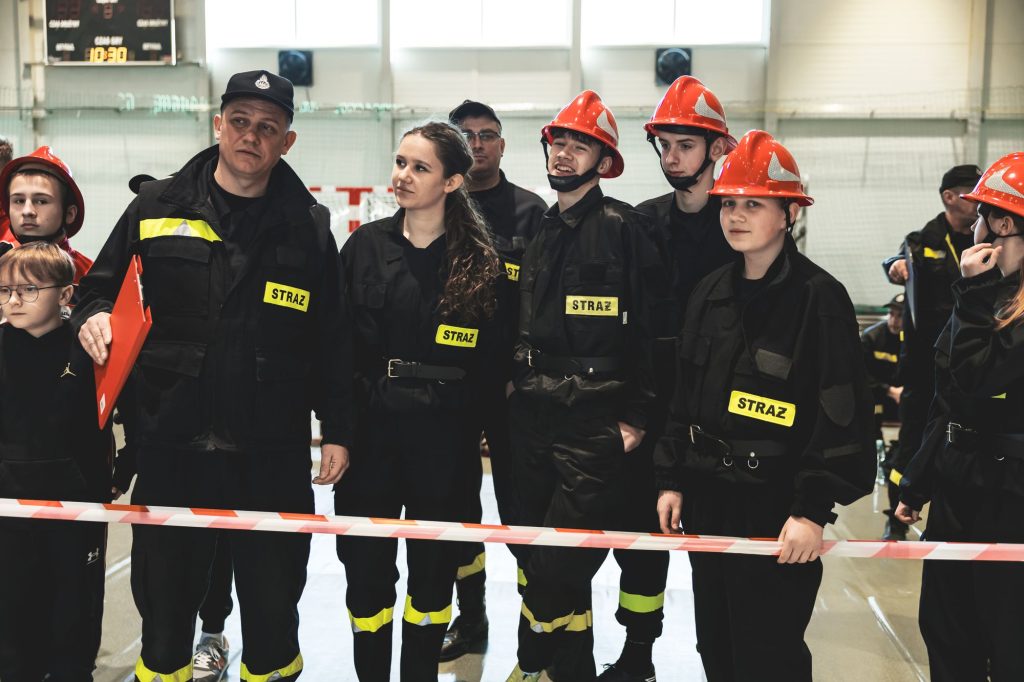 Grupa osób w czarnych mundurach z żółtymi naszywkami "STRAŻ", część w czerwonych hełmach, stojąca za białoczerwoną taśmą w jasnym pomieszczeniu.