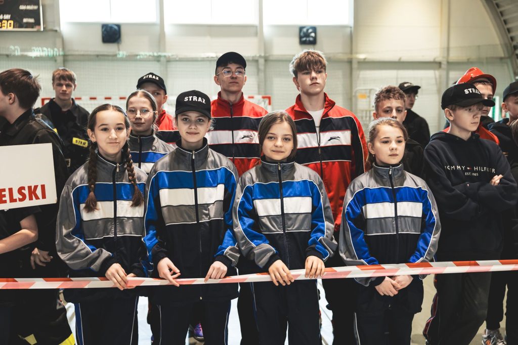 Grupa nastolatków w sportowych dresach w odcieniach niebieskiego, szarości i czerwieni, niektórzy w czapkach z napisem "STRAŻ", stoją za taśmą ostrzegawczą w hali.