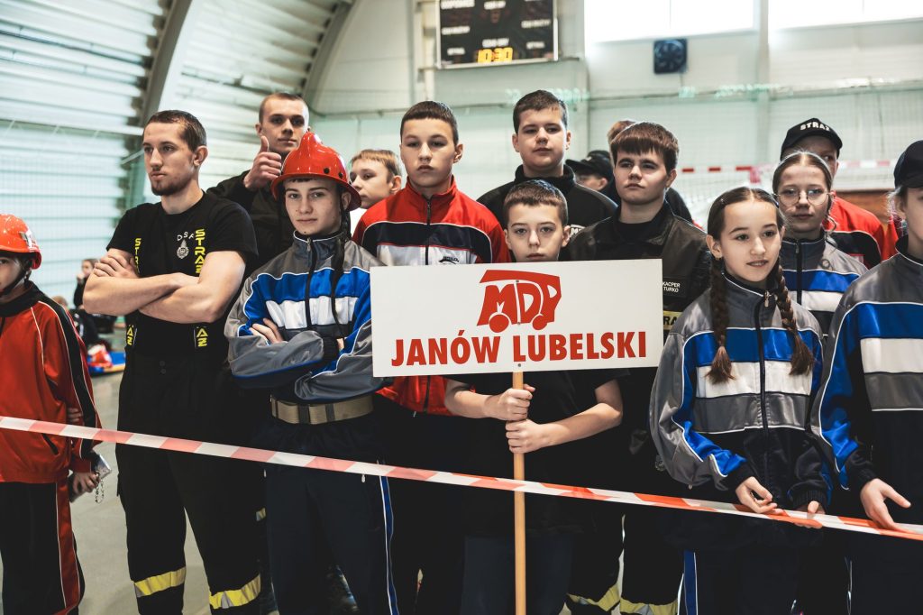 Grupa nastolatków w sportowych strojach, niektórzy w czerwonych hełmach, trzymający tablicę z napisem "MDP Janów Lubelski" w hali sportowej.
