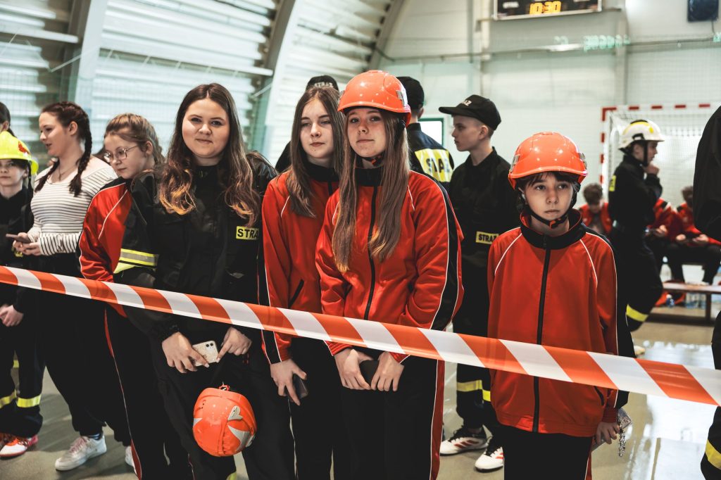 Grupa młodych osób w czerwonych i czarnych mundurach strażackich, niektóre w pomarańczowych hełmach, stoją za biało-czerwonym paskiem w hali sportowej.