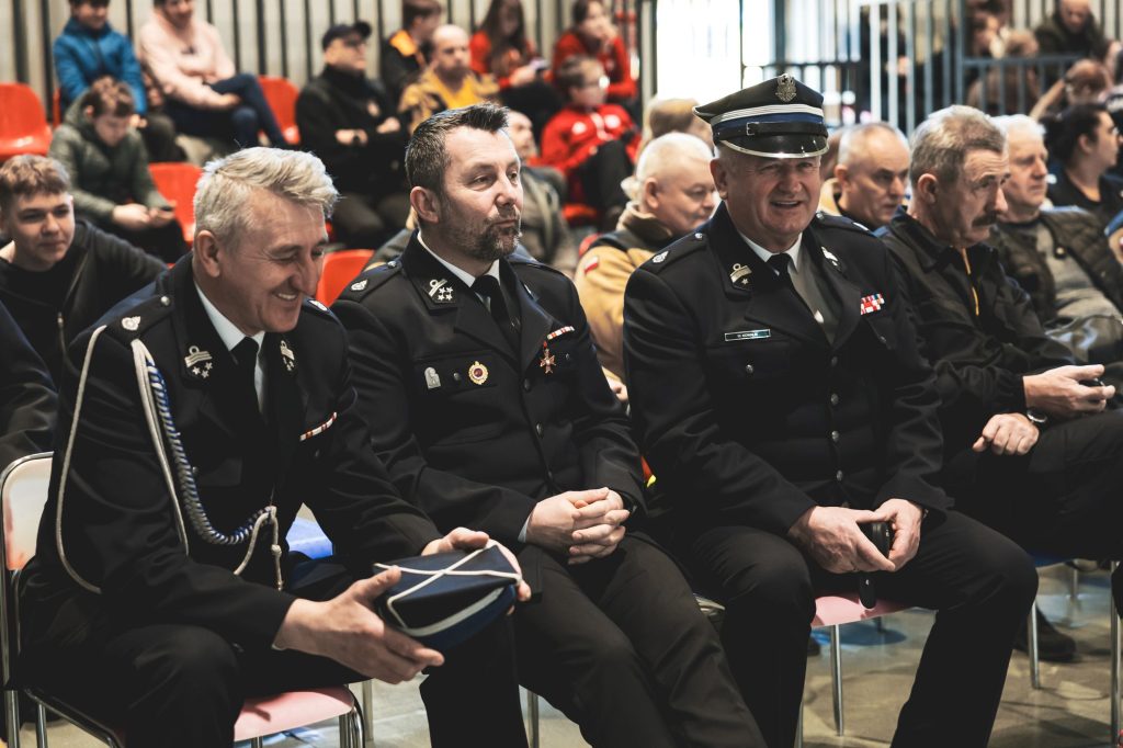 Trzech mężczyzn w mundurach z odznaczeniami siedzi na krzesłach w hali, za nimi ludzie na czerwonych trybunach.