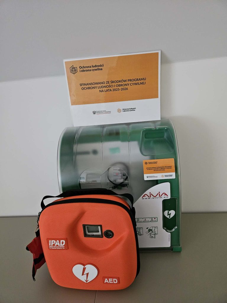 Pomarańczowa torba AED z symbolem serca i pioruna, zielona obudowa automatycznego defibrylatora, tablica z napisem o dofinansowaniu programu ochrony ludności na białym tle.