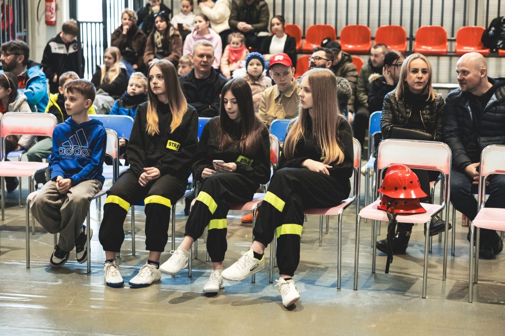 Grupa ludzi siedząca na metalowych krzesłach, trzy dziewczyny w czarnych mundurach z żółtymi odblaskami i napisami "STRAŻ", czerwone kaski na krześle, w tle ludzie na trybunach.