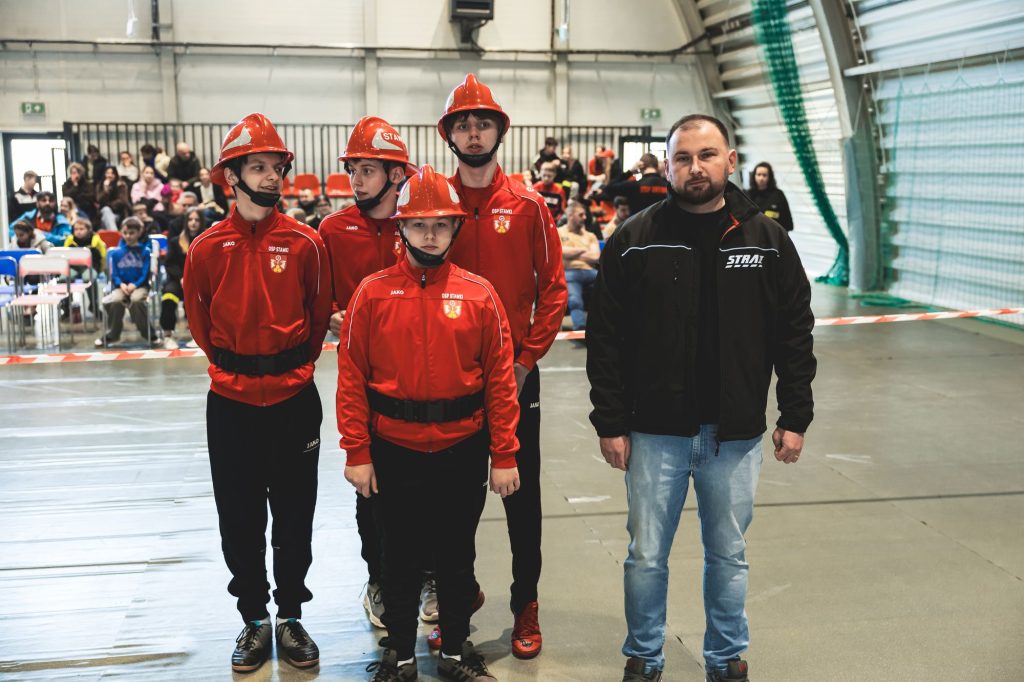 Czterech chłopców w czerwonych kurtkach i hełmach oraz mężczyzna w czarnej kurtce i jasnych jeansach na tle hali sportowej z widownią.