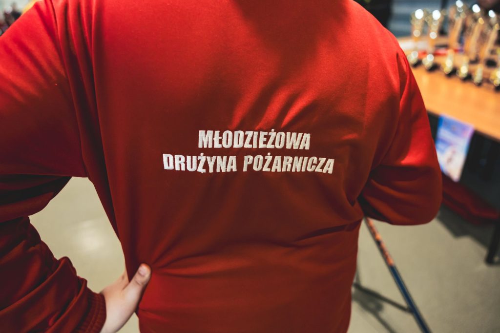 Czerwony strój z białym napisem "Młodzieżowa Drużyna Pożarnicza" na plecach, ręka podtrzymująca bok, w tle rozmyte trofea i kij.