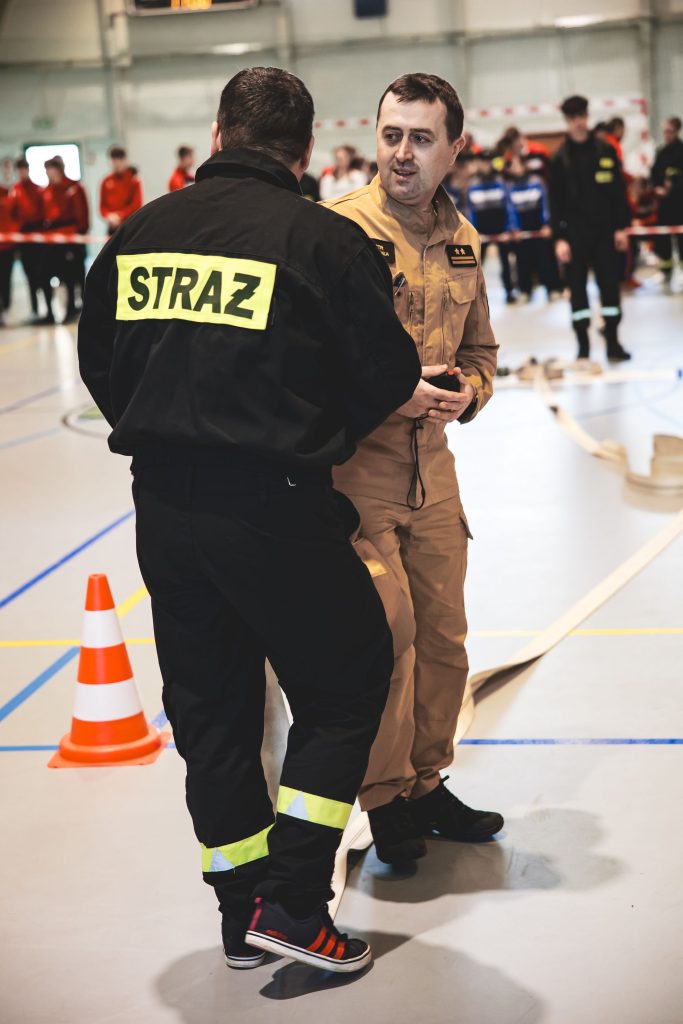 Dwaj mężczyźni w mundurach strażackich rozmawiają na hali sportowej z rozłożonymi wężami strażackimi i pachołkiem drogowym.
