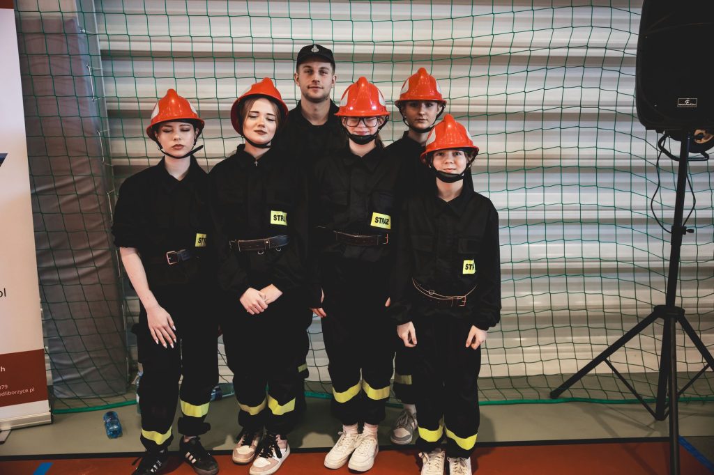 Grupa sześciu osób w czarnych mundurach z żółtymi naszywkami "STRAŻ" i czerwonych hełmach stoi na tle zielonej siatki.