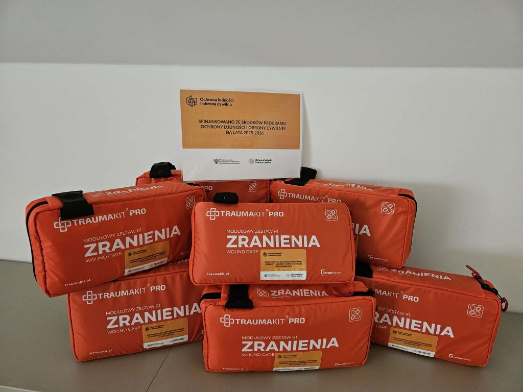 Sześć pomarańczowych zestawów pierwszej pomocy Traumakit Pro z napisem "Zranienia" ułożonych na szarym stole, na białym tle kartka z napisem o dofinansowaniu.