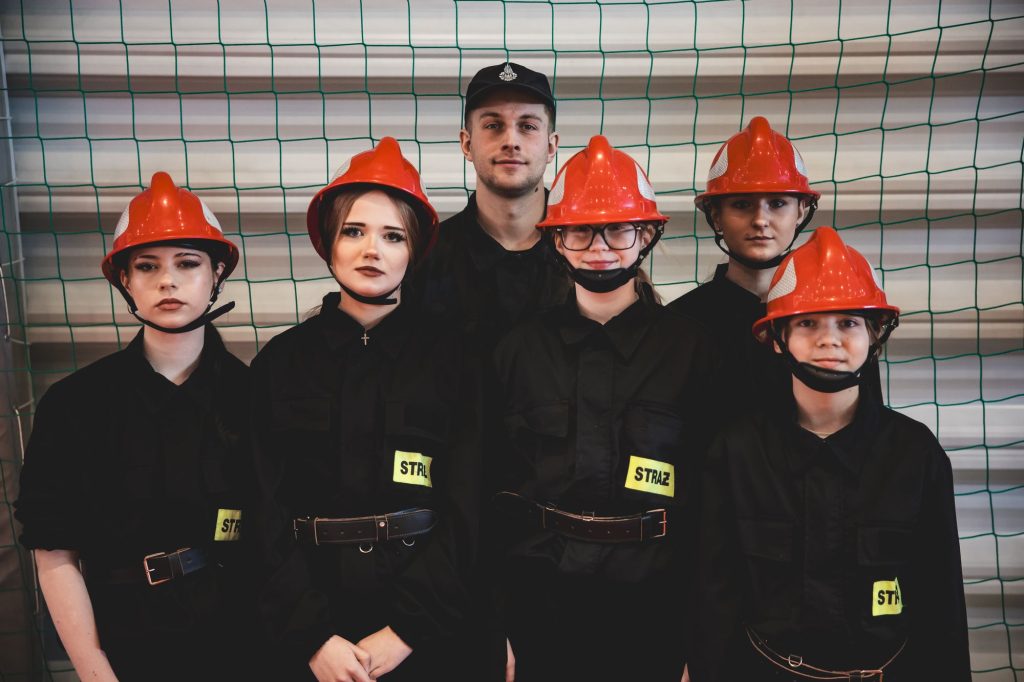 Grupa pięciu młodych osób w czarnych mundurach z żółtymi naszywkami "STRAŻ" i czerwonych hełmach, jedna osoba w czarnym czapce, w tle zielona siatka i jasna ściana.