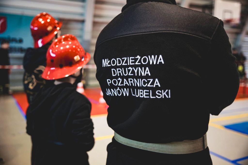 Czarna bluza z białym napisem "Młodzieżowa Drużyna Pożarnicza Janów Lubelski", dwie osoby w czerwonych kaskach, wnętrze hali sportowej z kolorowymi liniami na podłodze.