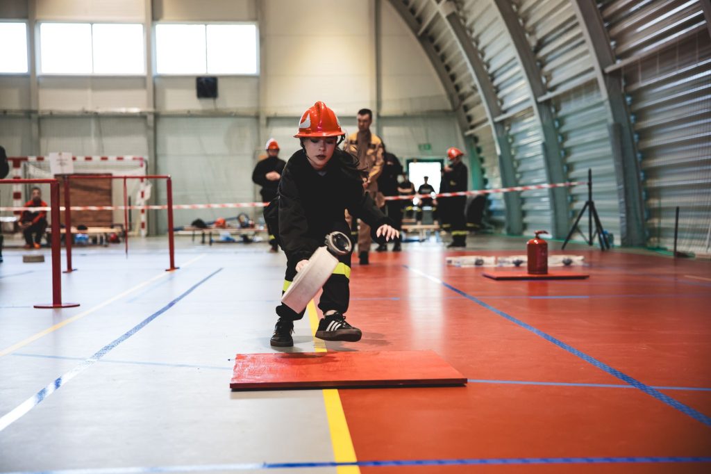 Kobieta w czerwonym hełmie i czarnym ubraniu z żółtymi paskami kuca na hali sportowej trzymając zwinięty wąż strażacki.