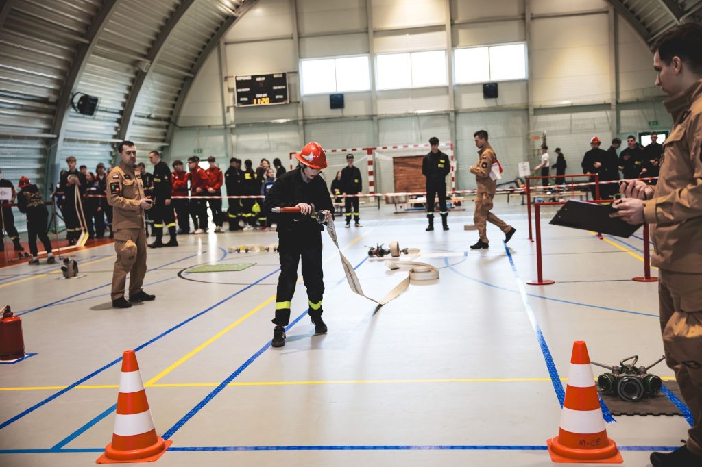 Osoba w czarnym ubraniu i czerwonym hełmie rozwija białą strażacką linię w hali sportowej, w tle ludzie w mundurach obserwują i notują, pomarańczowe pachołki na podłodze.