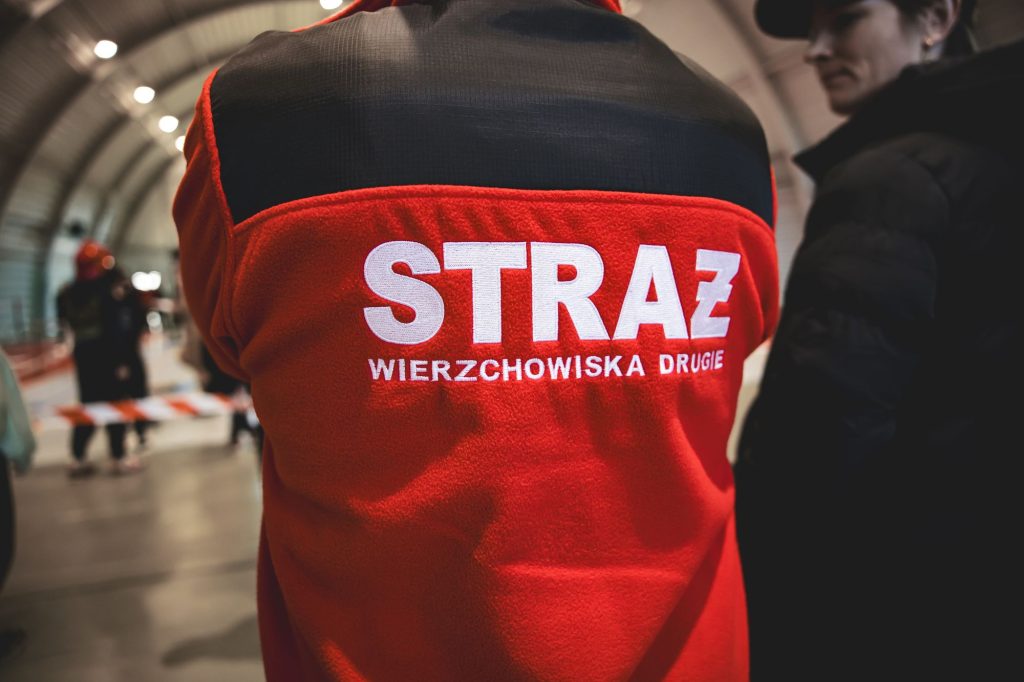 Czerwony polar z czarnym pasem u góry, na plecach biały napis STRAŻ WIERZCHOWISKA DRUGIE, w tle rozmyte postacie i wnętrze hali.
