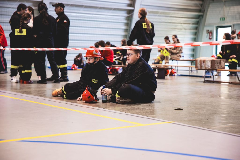 Dzieci w czarnych mundurach strażackich, czerwone hełmy, siedzą na podłodze hali sportowej, w tle grupa osób w podobnych mundurach przy ławkach i taśma ostrzegawcza.