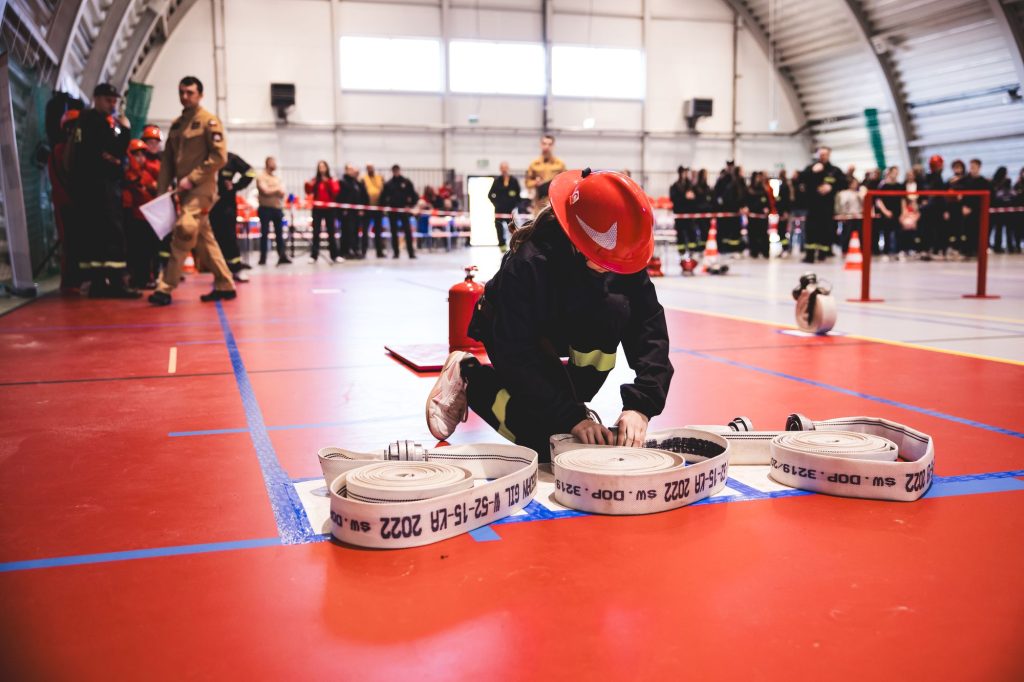 Osoba w czarnym ubraniu i czerwonym kasku zwija białe węże strażackie na czerwonej podłodze hali sportowej, w tle grupa ludzi i strażaków.