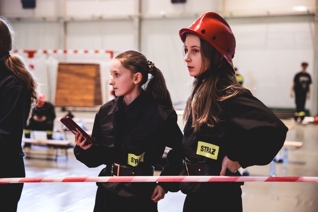 Dwie dziewczynki w czarnych mundurach strażackich z żółtymi naszywkami "STRAŻ", jedna z nich ma czerwony hełm, stoją za biało-czerwoną taśmą w hali sportowej.