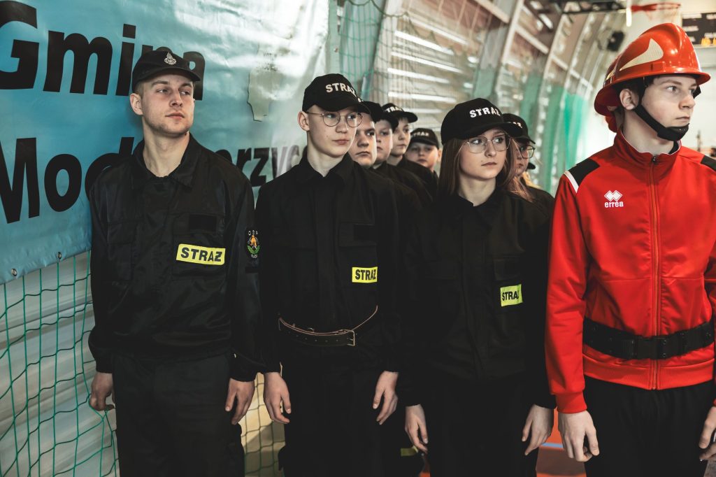 Grupa młodych strażaków w czarnych mundurach i czapkach z napisem "STRAŻ" stoi obok mężczyzny w czerwonej kurtce i czerwonym hełmie. W tle niebieska siatka i baner.