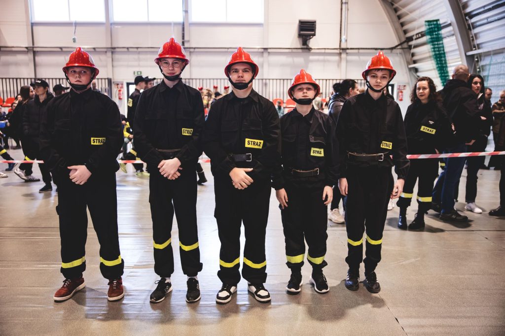 Pięciu młodych chłopców w czarnych mundurach z żółtymi napisami "STRAŻ" i czerwonych hełmach stoi w rzędzie w hali sportowej. W tle ludzie i barierka.