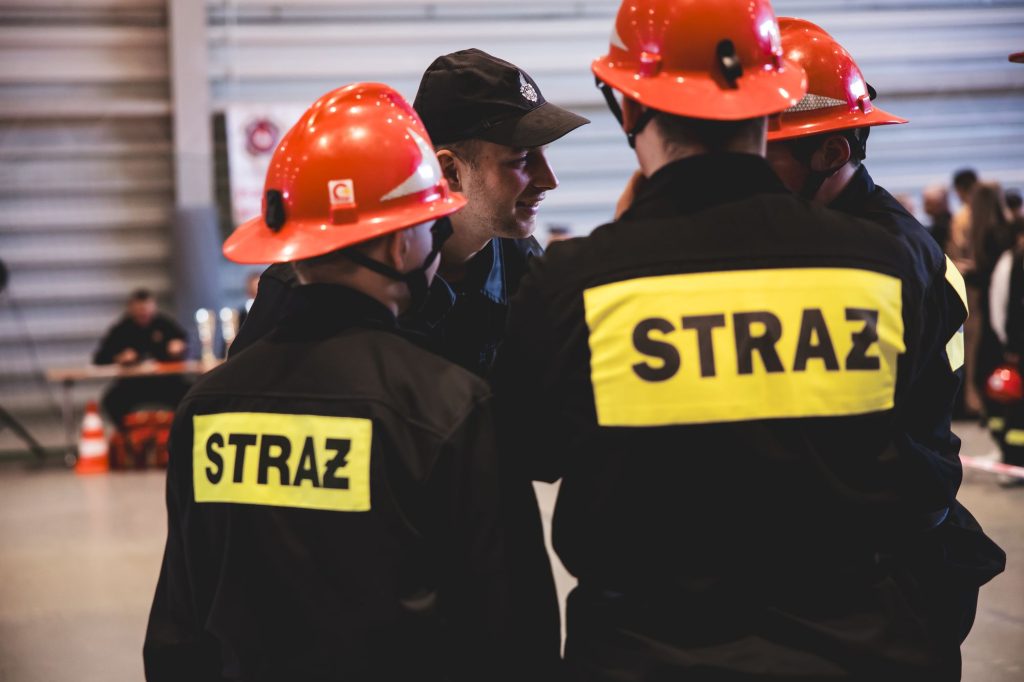 Kilka osób w czarnych mundurach z żółtymi napisami "STRAŻ" i czerwonych hełmach, rozmowa w hali z jasnymi ścianami i rozmytym tłem.
