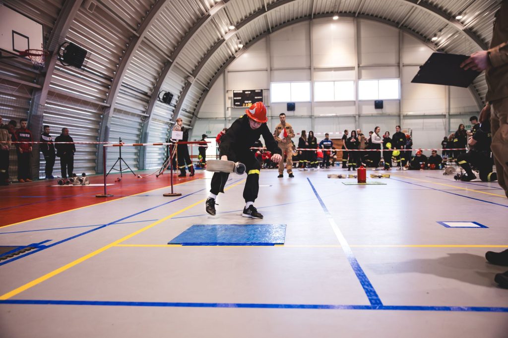 Osoba w czarnym ubraniu i czerwonym hełmie w hali sportowej trzyma zwinięty wąż strażacki, w tle ludzie i sprzęt strażacki.
