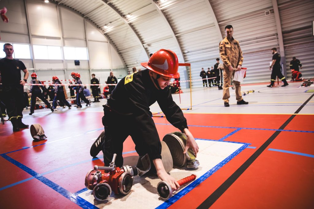 Osoba w czarnym ubraniu i czerwonym hełmie układa zwinięte węże strażackie na jasnej macie na czerwonej podłodze hali sportowej. W tle inni ludzie i sprzęt.