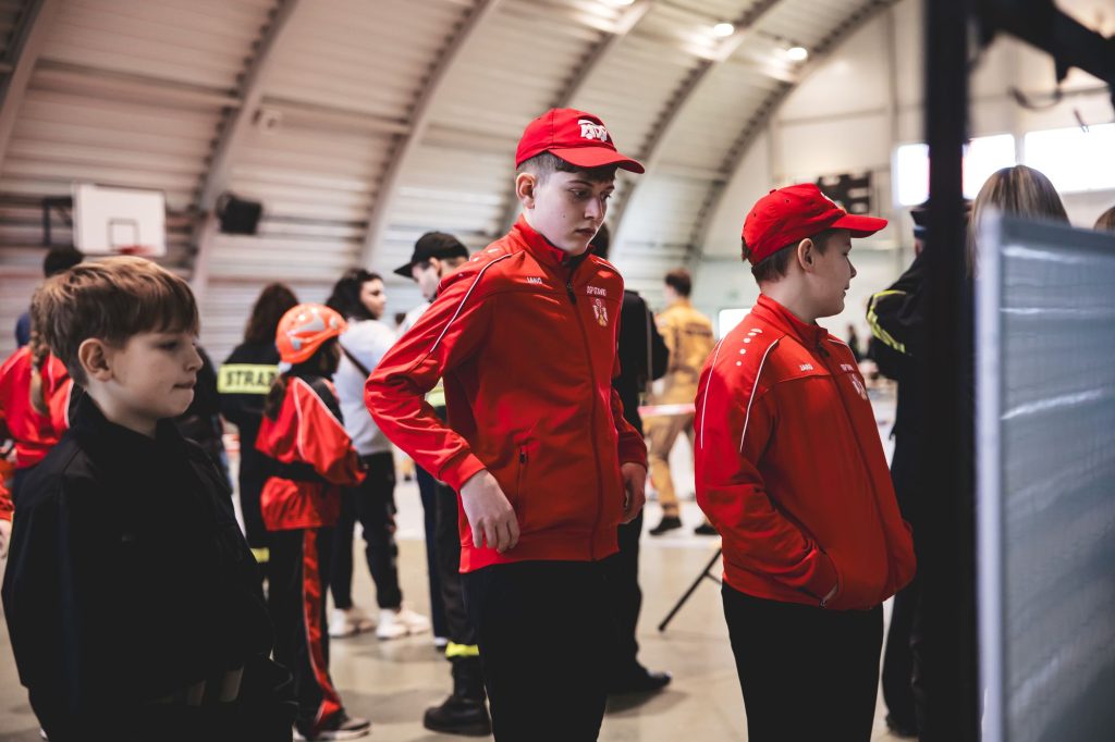 Dwóch chłopców w czerwonych kurtkach i czapkach, obok nich chłopiec w czarnej bluzie, w tle osoby w strojach roboczych i kasku, hala sportowa.