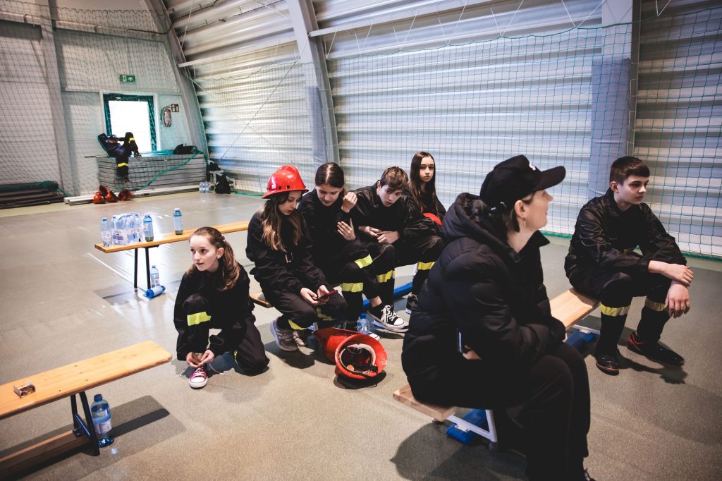 Grupa młodzieży w czarnych kombinezonach z żółtymi paskami siedzi i klęczy na drewnianych ławkach w hali sportowej z siatką na ścianie.